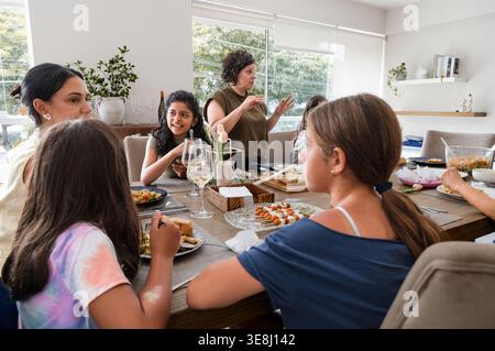 Une famille hispanique multigénérationnelle assise autour d'une table à manger en bois profitant d'un repas ensemble. Les femmes et les filles s'engagent dans la conversation et e Banque D'Images