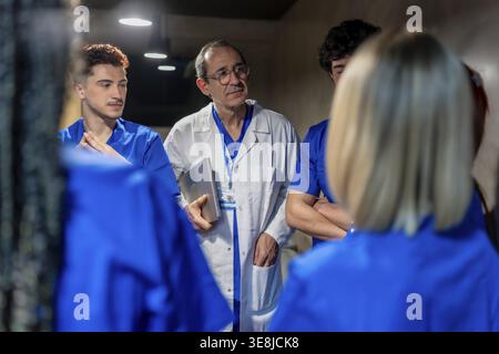 Médecin masculin expérimenté portant une blouse de laboratoire et tenant un ordinateur portable, s'engageant avec un groupe de jeunes étudiants en médecine dans des gommages bleus pendant un hôpital educ Banque D'Images