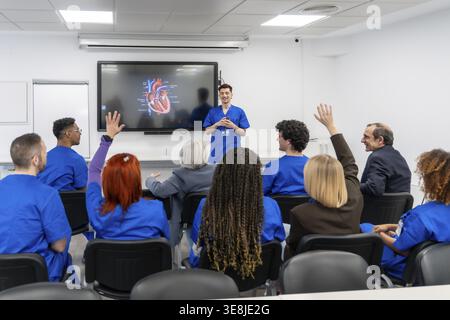 Un groupe diversifié d'étudiants et de professionnels de la santé participent activement à un séminaire de formation médicale, levant la main pour poser des questions lors d'une pré Banque D'Images