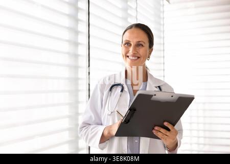 Médecin féminin souriant tenant une planche à pince à la clinique Banque D'Images