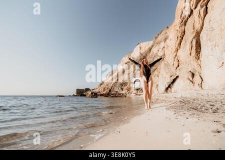 Femme plage vacances femme heureuse en maillot de bain noir marche sur une plage de sable avec les bras ouverts large par les falaises de l'océan Banque D'Images