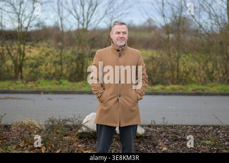 Un homme âgé confiant portant un manteau marron se tient dehors avec ses mains dans ses poches tout en regardant calmement au loin. Le backgrou naturel Banque D'Images