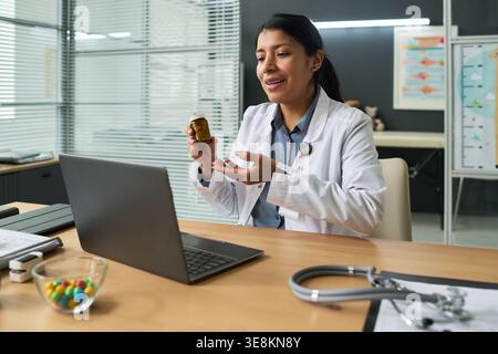 Plan moyen d'un jeune médecin adulte latin tenant une bouteille de médicament et expliquant la posologie pendant la consultation avec un ordinateur portable. Soutien à la télésanté, pharmacie en ligne Banque D'Images