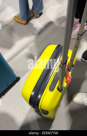 Vue en angle élevé d'une valise jaune vif avec des roues et une poignée télescopique tenue par un voyageur dans un terminal d'aéroport ou une gare très fréquentée Banque D'Images