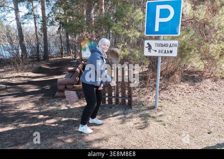 Chelyabinsk, Russie - 2 avril 2026 : femme âgée joyeuse jouant avec des sorcières humoristiques seul signe de stationnement, tenant un balai dans la forêt ensoleillée Banque D'Images