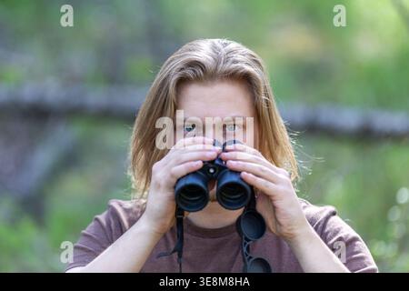 Portrait en gros plan d'un jeune homme aux longs cheveux blonds et aux yeux bleu clair. Il tient une paire de jumelles mais regarde directement la caméra au-dessus d'elles. Banque D'Images