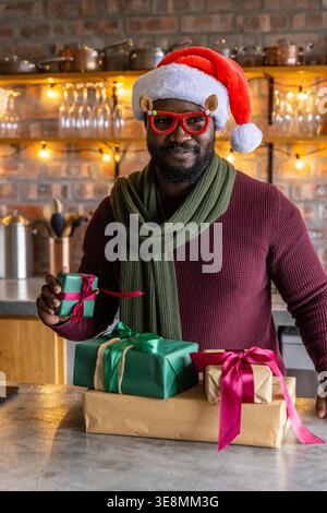 Homme afro-américain portant le chapeau de Père Noël et les spécifications de nouveauté tenant le cadeau vert dans la cuisine chaude. Festif, confortable, brique, comptoir, stringlights, bourgogne, écharpe Banque D'Images