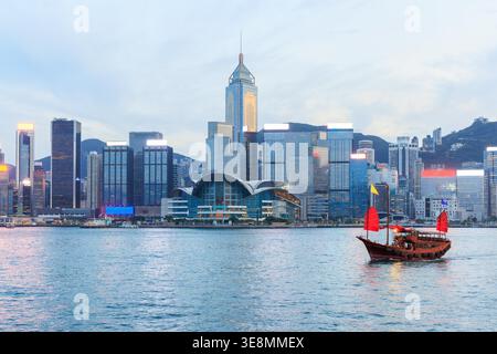 Jonque traditionnelle en bois avec des voiles rouges contre Hong Kong skyline pendant le coucher du soleil, scène urbaine emblématique Banque D'Images