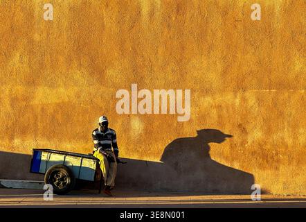 Vendeur de rue se reposant à côté d'un chariot jetant une longue ombre sur un mur ensoleillé à Essaouira, Maroc Banque D'Images