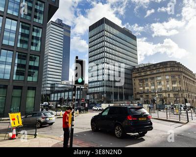 Manchester, Royaume-Uni - 22 février 2026 : rue animée de Manchester UK avec tours de bureaux, voitures, feux de circulation et un piéton à l'intersection. Banque D'Images