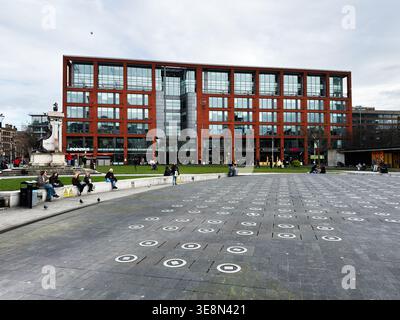 Manchester, Royaume-Uni - 22 février 2026 : Urban plaza à Manchester, Royaume-Uni avec un immeuble de bureaux en briques rouges, des gens assis et marchant. Banque D'Images
