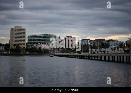 Divers bâtiments le long de la rive de St Kilda Beach, vus de l'autre côté de l'eau avec St Kilda Pier au premier plan, lors d'une soirée sombre Banque D'Images