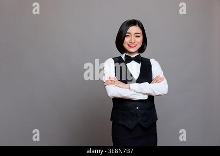 Souriante serveuse asiatique confiante en uniforme debout avec le portrait des bras croisés. Jeune réceptionniste professionnelle joyeuse posant les mains pliées et regardant la caméra Banque D'Images