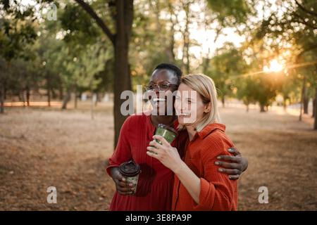 Deux heureux amis multi-ethniques riant et serrant dans un parc au coucher du soleil, buvant du café Banque D'Images