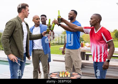 Divers amis masculins grillent avec des bouteilles vertes après l'homme prolongeant la bouteille tout en grillant au gril Banque D'Images