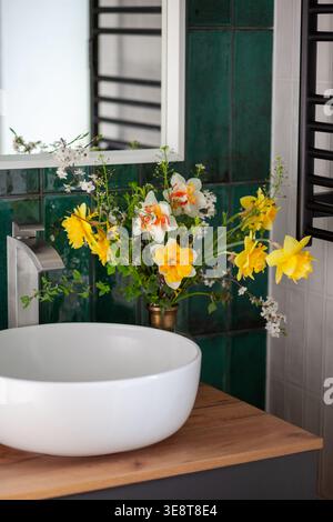 Intérieur de salle de bain confortable avec bouquet de fleurs de printemps dans un vase. Conception moderne du bassin rond sur l'unité de vanité par mur carrelé vert Banque D'Images