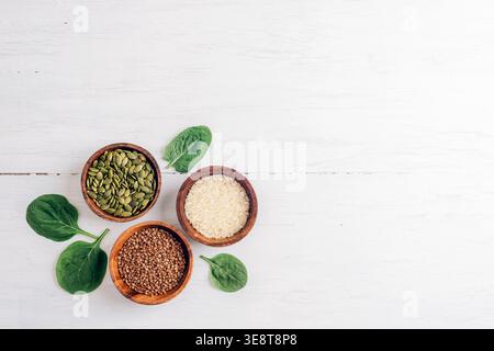 Céréales et graines dans des bols et des feuilles vertes sur une table en bois blanc. Concept de nourriture saine. Riz, sarrasin et graines de citrouille. Vue de dessus, pose à plat, copie s Banque D'Images