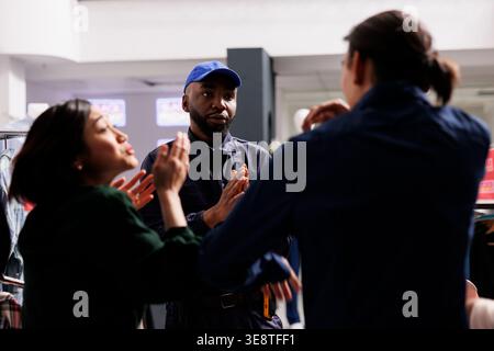 Calme poli Afro-américain homme agent de sécurité gérant la foule dans le centre commercial pendant les ventes, demandant à deux acheteurs fous en colère de rester calme pendant le Black Friday. Les gens marchandent les chasseurs en combattant Banque D'Images
