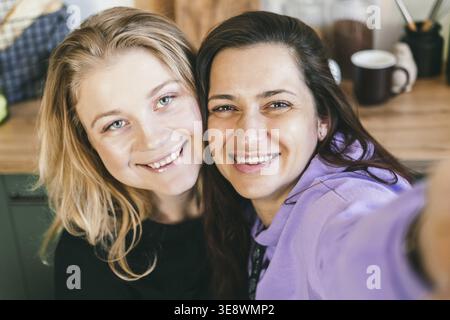 Les copines rient joyeusement et prennent des selfies dans la cuisine. Deux filles sont photographiées au téléphone. Brunette et blonde s'amusent. Les femmes regardant sma Banque D'Images