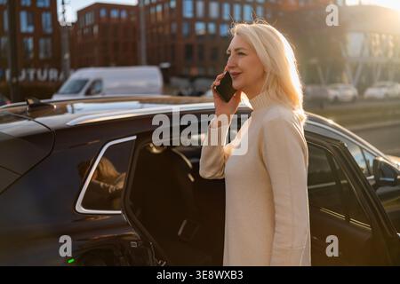 Une femme professionnelle s'engage dans une conversation téléphonique en sortant d'une berline noire élégante. L'environnement urbain comprend des bâtiments modernes et une lumière douce du soleil illuminant la scène. Banque D'Images