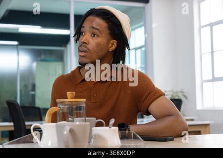 Homme afro-américain 20s portant le polo de rouille de chapeau en tricot assis dans le bureau versant de la théière en verre Banque D'Images