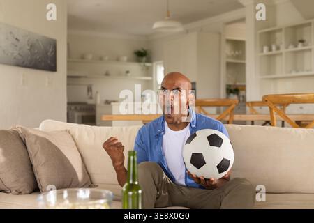 Homme afro-américain assis sur le canapé tenant le ballon de football, criant, bouteille et pop-corn à proximité Banque D'Images
