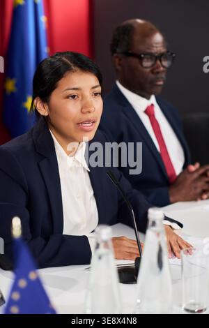 Plan vertical d'une jeune femme adulte latine prenant la parole lors d'une conférence de presse aux côtés d'un homme noir d'âge moyen, transmettant la communication du gouvernement et le leadership Banque D'Images
