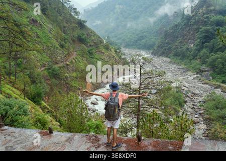 Un homme se tient sur une falaise surplombant une vallée sereine avec une rivière qui coule à travers elle au temple Madhyamaheshwar, Uttarakhand, Inde. La vue panoramique au large Banque D'Images