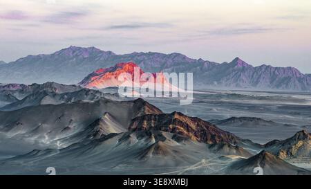 Lumière du coucher de soleil illuminant la montagne Yanzhi au-delà des crêtes sombres de la montagne Heidu dans le Qinghai, en Chine. La perspective aérienne capture des contrastes spectaculaires. Banque D'Images