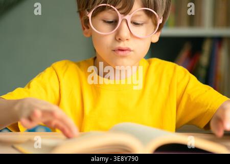 Élève souriant dans des lunettes repose la main tout en lisant un livre ouvert au bureau de l'école. Apprentissage de l'enfance, esprit intelligent, concept d'étude, développement précoce et préparation à l'éducation et à la croissance future. Banque D'Images