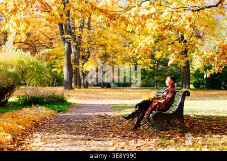Jolie femme européenne rousse en manteau à carreaux et béret noir assis sur un banc dans le parc d'automne à la journée ensoleillée, vue de côté. Humeur d'automne, concept de temps de loisirs. Temps de détente dans le parc de la ville Banque D'Images