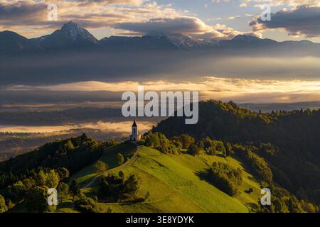 Vue aérienne de l'église de préparus Primus et Felician sur une crête herbeuse surplombant une vallée brumeuse et les Alpes Kamnik-Savinja pendant le lever du soleil Jamnik, K Banque D'Images
