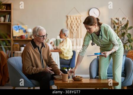Homme âgé recevant de l'eau d'une jeune infirmière en maison de retraite, soutenant les soins quotidiens et l'assistance à la vie Banque D'Images