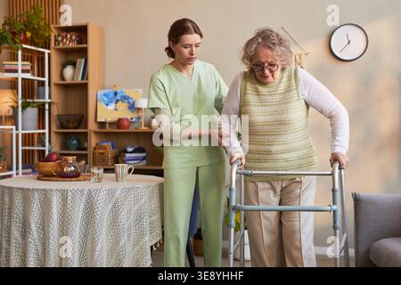 Femme âgée utilisant un marcheur avec une aidante soignante pendant la pratique de la mobilité en maison de retraite Banque D'Images