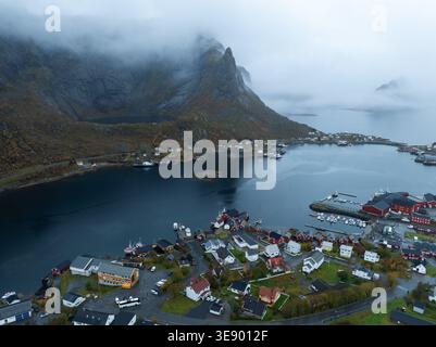 Vue aérienne du pittoresque village de Reine avec ses maisons rouges traditionnelles nichées entre les eaux calmes du fjord et les montagnes couvertes de brume à Reine, Banque D'Images