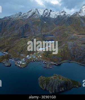 Vue aérienne du village de pêcheurs de Nusfjord avec des cabanes rouges traditionnelles, entouré de montagnes escarpées enneigées et des eaux sombres du fjord Nusfjord, Nordla Banque D'Images
