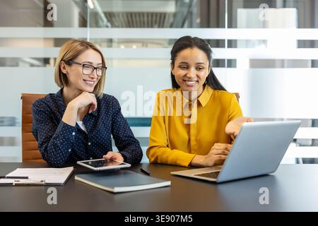 Deux femmes d'affaires souriantes et collaborant sur un ordinateur portable et une tablette dans un bureau moderne lumineux, examinant les données du projet et la stratégie de planification ensemble dans un cadre convivial et professionnel Banque D'Images
