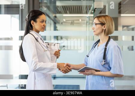 Deux professionnelles médicales féminines, un médecin en blouse de laboratoire et une infirmière en gommage, scellant un partenariat professionnel en serrant la main dans un environnement de soins de santé moderne Banque D'Images