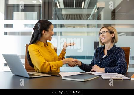 Deux femmes d'affaires sourient et serrent la main sur un bureau avec un ordinateur portable et des documents, symbolisant un partenariat, une collaboration et un accord réussis Banque D'Images