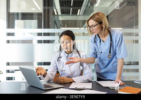 Deux femmes médecins collaborant et analysant des documents médicaux et des informations sur un ordinateur portable lors d'une discussion sérieuse dans un bureau moderne ou un cadre clinique Banque D'Images