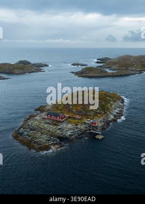 Vue aérienne d'une cabane traditionnelle en bois rouge sur une petite île rocheuse entourée par les eaux bleues profondes de l'archipel des Lofoten sous une humeur changeante Banque D'Images