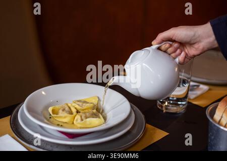Incliner à la main une théière en céramique blanche verser un bouillon clair sur une assiette blanche profonde contenant de grands tortellini jaunes. Table sombre avec verre à eau, pain, a Banque D'Images
