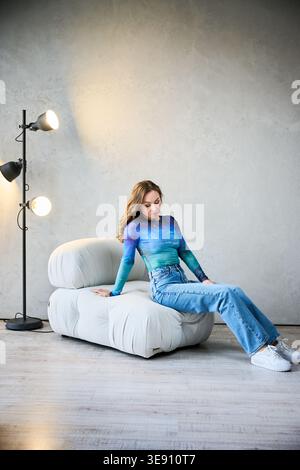 Femme gracieuse en haut artistique et jeans se trouve confortablement sur une chaise en peluche dans un espace contemporain. La lumière douce de la lampe projette une lueur chaleureuse, améliorant l'atmosphère chaleureuse idéale pour la détente. Banque D'Images
