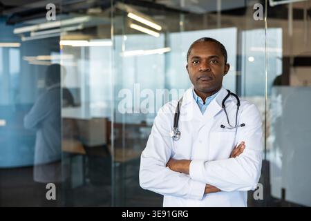 Médecin masculin expérimenté debout avec confiance avec les bras croisés, portant une blouse de laboratoire blanche et un stéthoscope, représentant le professionnalisme de la santé et l'expertise médicale dans un cadre de bureau Banque D'Images