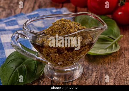 Gros plan sur une sauce pesto vert éclatante dans une saucière en verre transparent. Des feuilles de basilic frais et des tomates rouge vif sont visibles en arrière-plan Banque D'Images