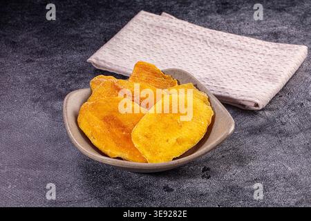 Tranches de mangue séchée orange vif dans un bol en céramique neutre. Disposé sur une surface texturée sombre avec une serviette pliée légère. Éclairage studio en surbrillance Banque D'Images