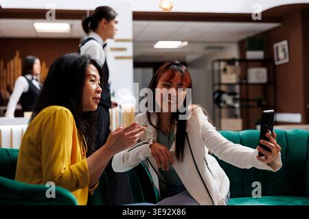 Heureux touristes asiatiques prenant des selfies dans le hall d'accueil de l'hôtel pendant les vacances. Deux clients arrivant en utilisant leur smartphone pour prendre des photos pendant leurs vacances dans le salon contemporain de l'hôtel Banque D'Images