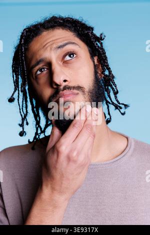 Jeune homme arabe se frottant le menton en contemplation avec une expression faciale perplexe. Modèle attrayant avec des tresses noires coiffure regardant loin tout en pensant avec des émotions pensives Banque D'Images