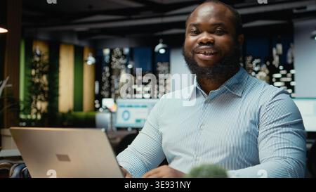 Portrait d'homme d'affaires noir fait la pensée critique et la relecture des fichiers avant de transcrire les données sur ordinateur portable, assis dans le bureau la nuit. Documenter les idées importantes heures supplémentaires. Banque D'Images