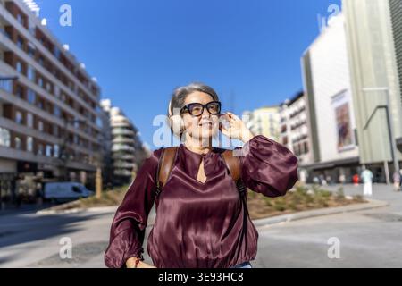Élégante femme d'âge moyen avec les cheveux gris et les lunettes se promène en toute confiance le long d'une rue ensoleillée de la ville, souriant et profitant de la musique à travers des écouteurs, détendez-vous Banque D'Images